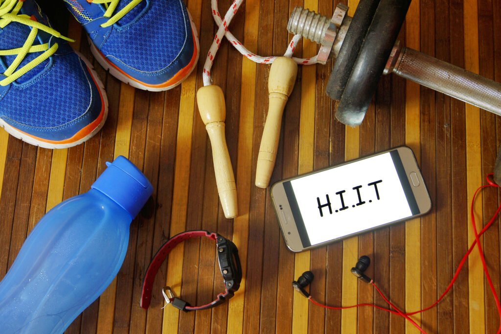 Hanteln, Sportschuhe, Smartphone mit Kopfhörern, Springseil und Wasserflasche auf dem Fitnessstudio-Boden mit der Aufschrift „H.I.I.T.“ (High Intensity Interval Training)