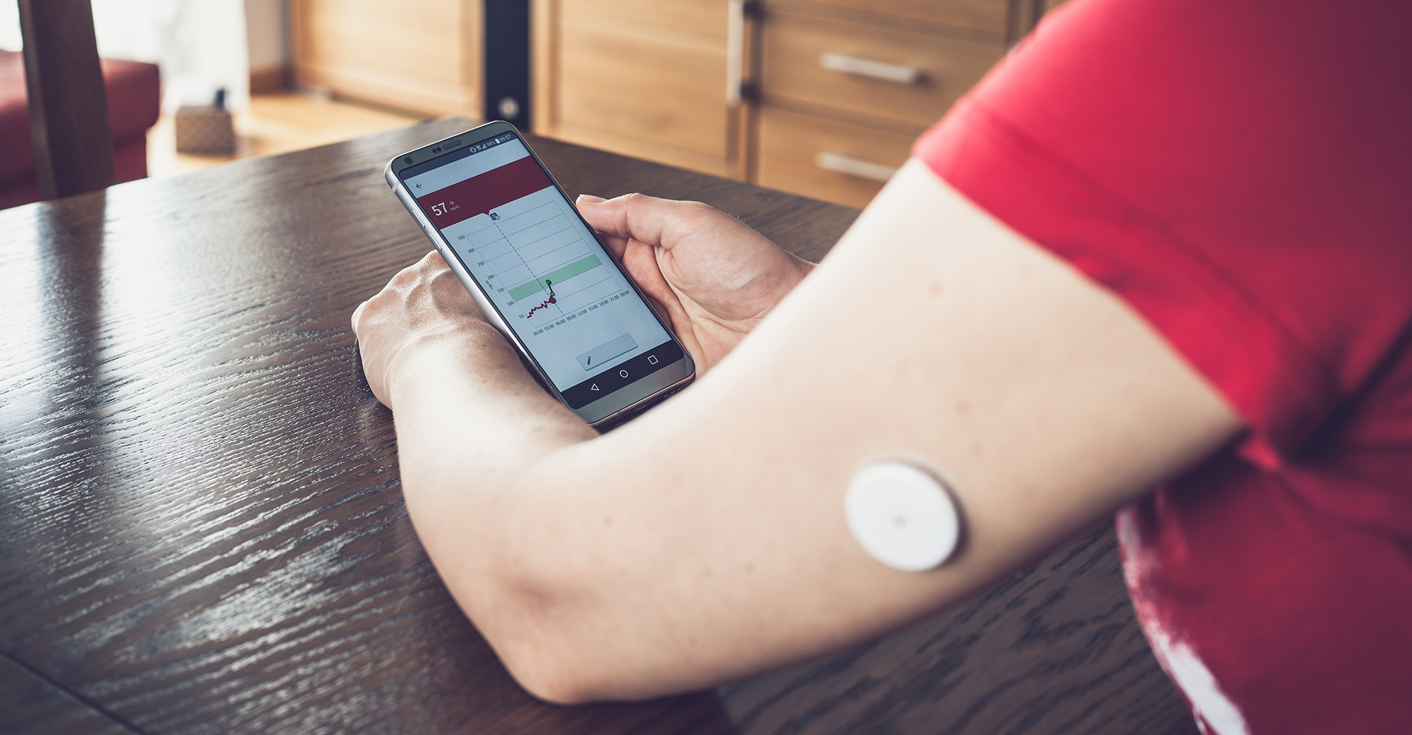 Frau sitzt an einem Holztisch und hält ein Smartphone in den Händen, am Oberarm trägt sie einen Sensor zur kontinuierlichen Glukosemessung.