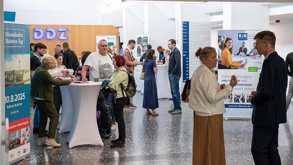 Belebte Informations- und Networking-Zone im Foyer einer Veranstaltung zum Thema Diabetes. Mehrere Personen stehen in kleinen Gruppen oder an Stehtischen verteilt, führen Gespräche oder informieren sich an Ständen.