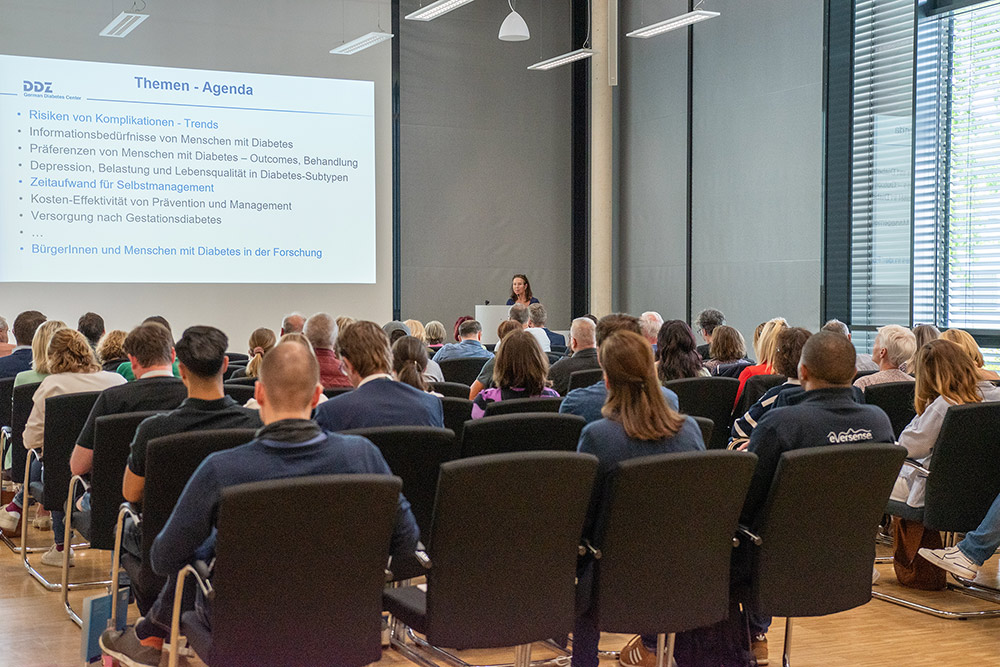 Bei einer Informationsveranstaltung in einem Konferenzraum steht eine Rednerin vorne am Podium und hält einen Vortrag vor dem Publikum.